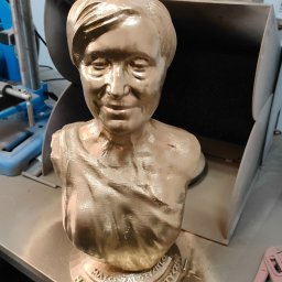 B3Ddesign.pl - Złoty, pomalowany biust osoby, wydrukowany w 3D, z inskrypcją na podstawie. W tle widoczny warsztat z narzędziami, co sugeruje proces produkcyjny.