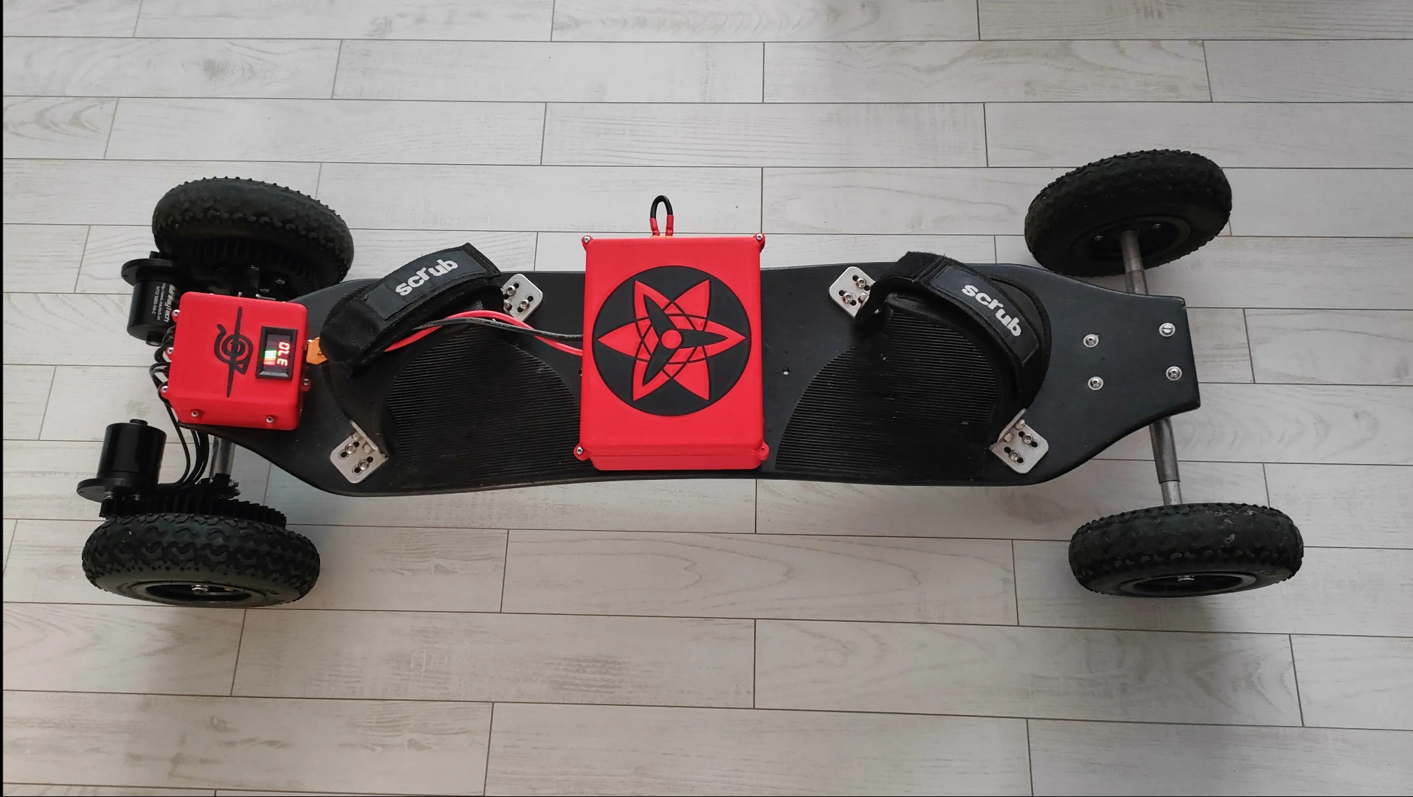 Elektryczna deska terenowa z napędem na cztery koła, widoczny czerwony moduł sterujący z wyświetlaczem i logo w kształcie gwiazdy w czarnym okręgu, leżąca na podłodze z paneli.