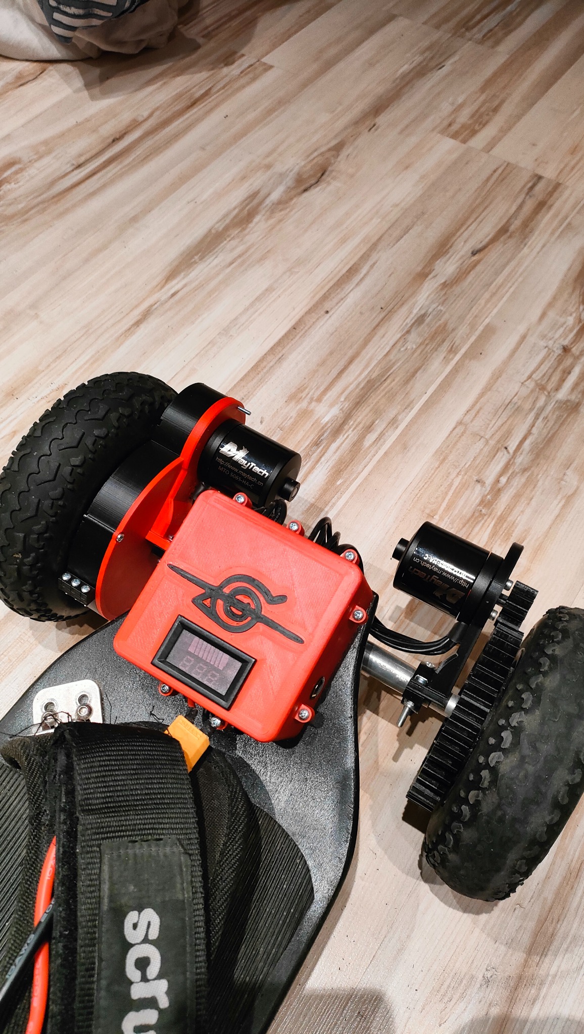 Elektryczna deska z napędem na dwa koła, widoczne silniki Maytech, czerwona obudowa elektroniki z wyświetlaczem i symbolem, zębatki napędowe, całość na tle paneli podłogowych.