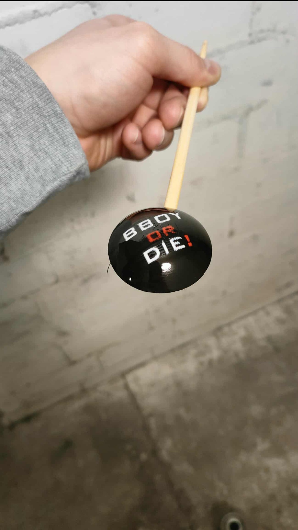 Ręka trzyma czarną, błyszczącą gałkę zmiany biegów z napisem 'Bboy or Die!' na drewnianym drążku, wykonaną prawdopodobnie metodą druku 3D.