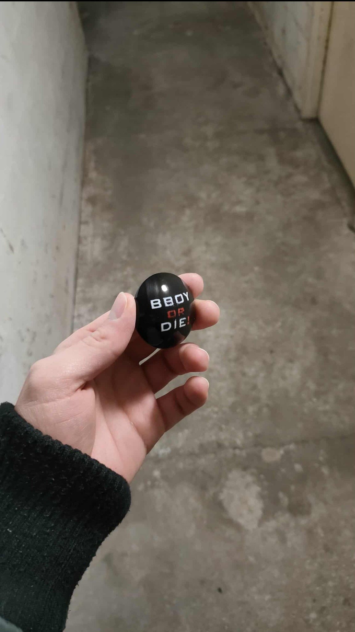 Dłoń trzyma czarną, okrągłą nakładkę na drążek zmiany biegów z białym napisem 'BBOY OR DIE!', wykonaną w technologii druku 3D FDM ASA.