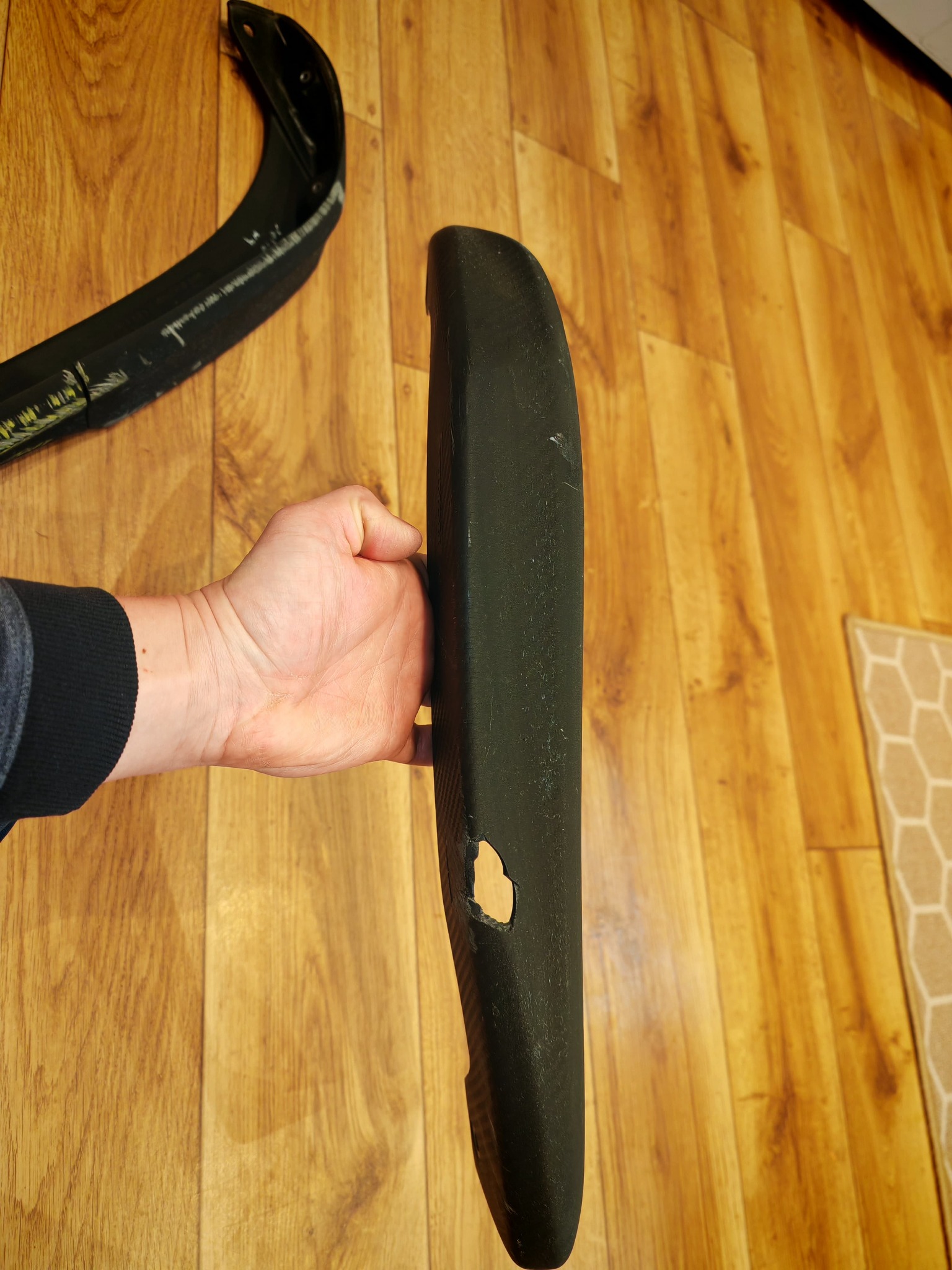 Czarny element z tworzywa sztucznego z otworem, trzymany w dłoni na tle drewnianej podłogi. Widoczny fragment drugiego elementu z tworzywa.