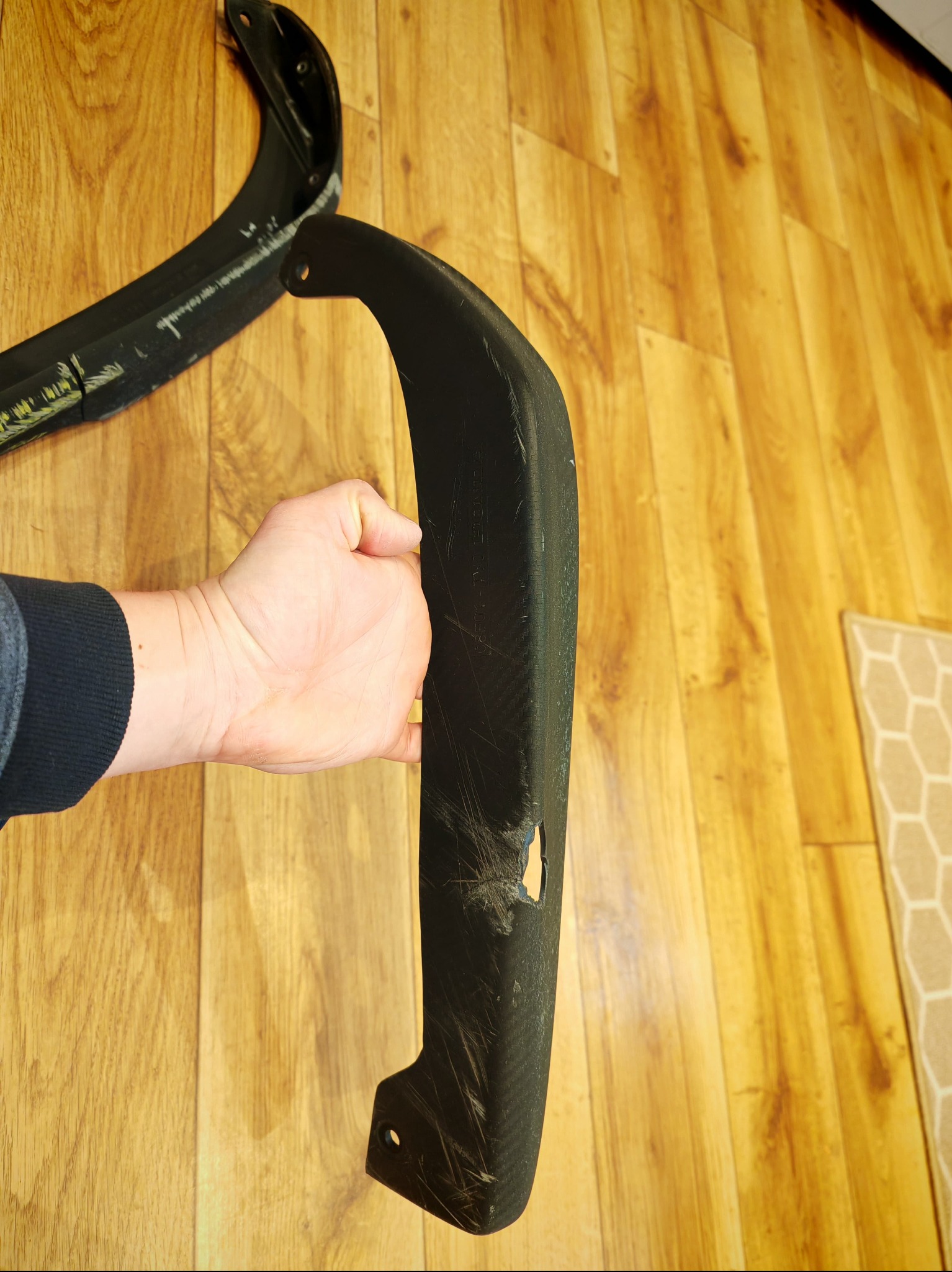 Czarny, zakrzywiony element z tworzywa sztucznego z otworem, trzymany w dłoni na tle drewnianej podłogi. Widoczne ślady użytkowania i uszkodzenia.