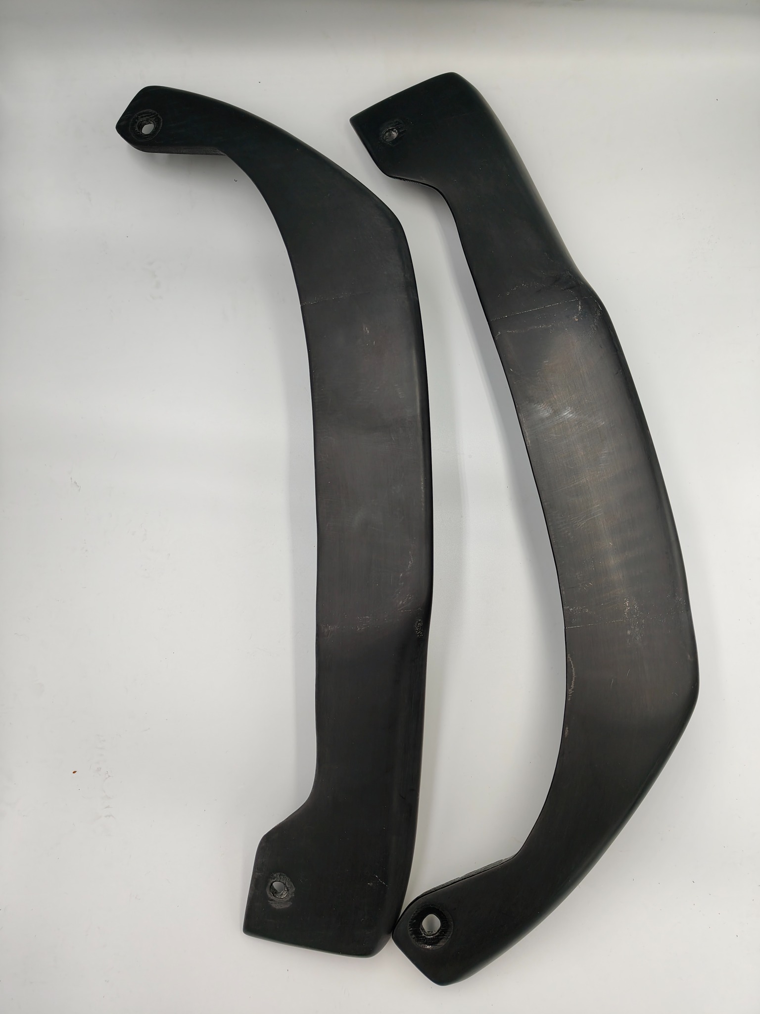 Dwa czarne, zakrzywione elementy z otworami montażowymi, prawdopodobnie wykonane z tworzywa sztucznego, leżące na białym tle. Widoczne ślady obróbki.