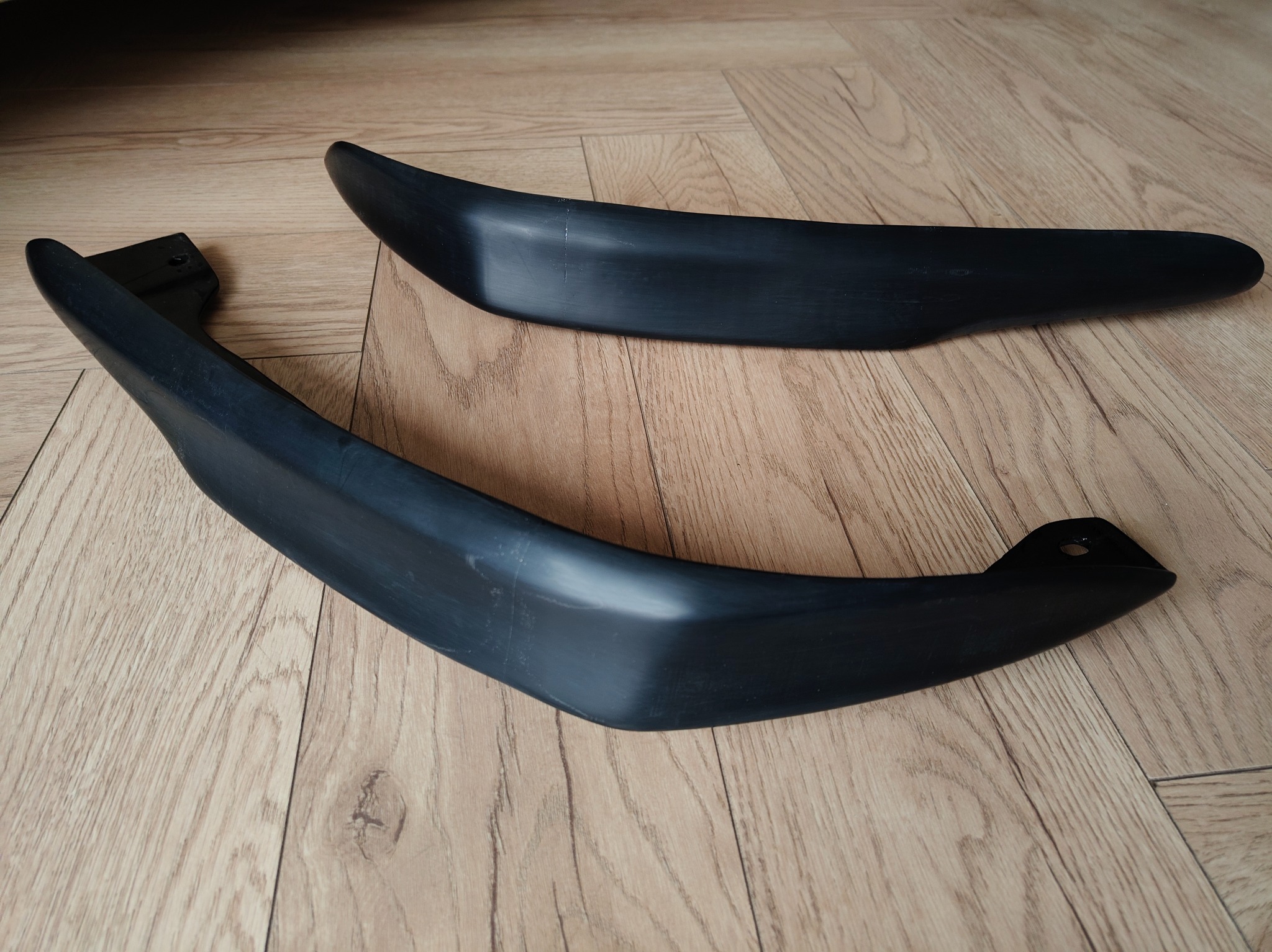Dwa czarne, matowe elementy z tworzywa sztucznego o zakrzywionym kształcie, leżące na panelach podłogowych. Jeden element z widocznym otworem montażowym.