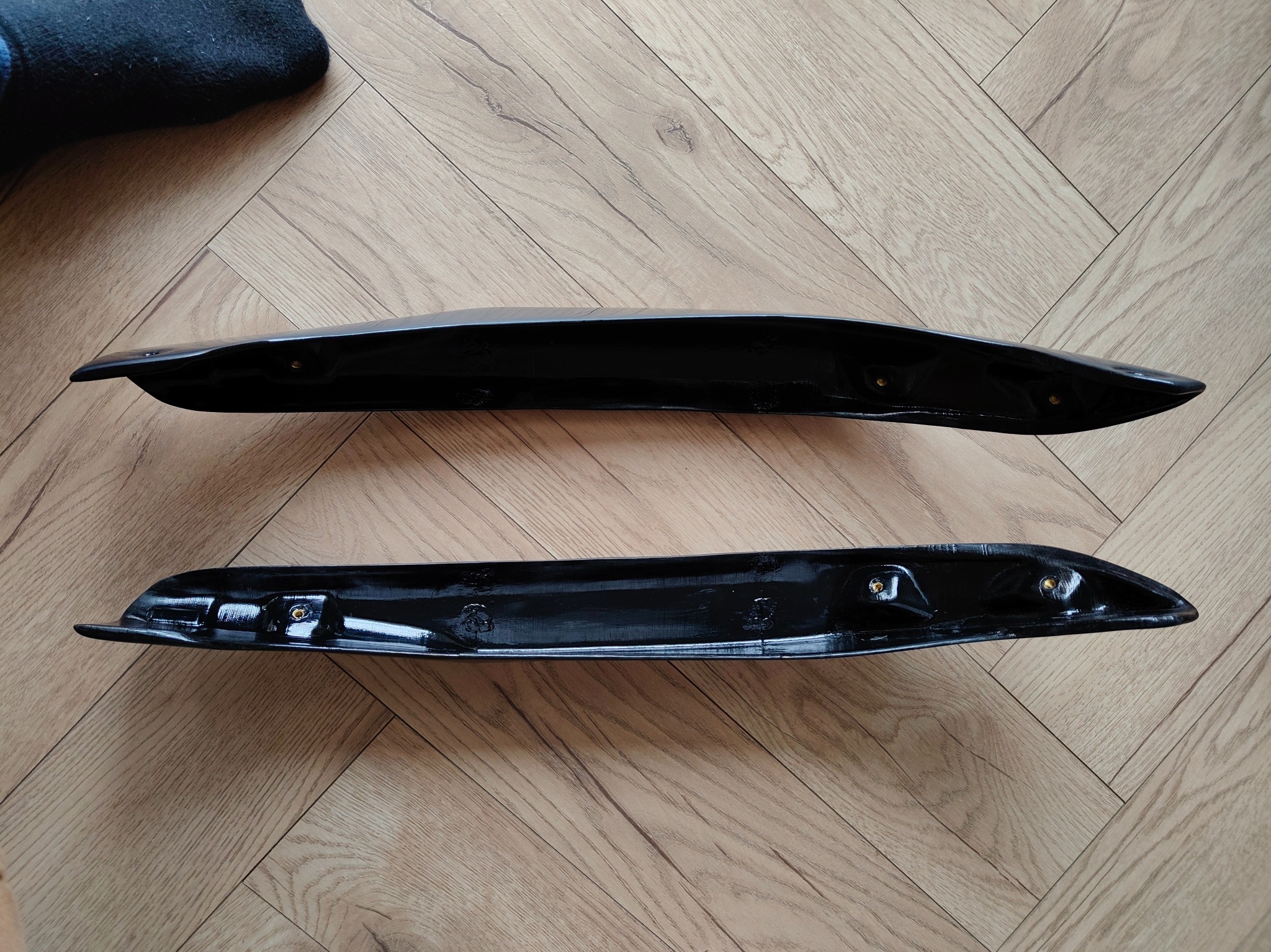 Dwie czarne, błyszczące osłony z tworzywa na drewnianej podłodze. Widoczne otwory montażowe i odbicia światła podkreślające gładką powierzchnię.