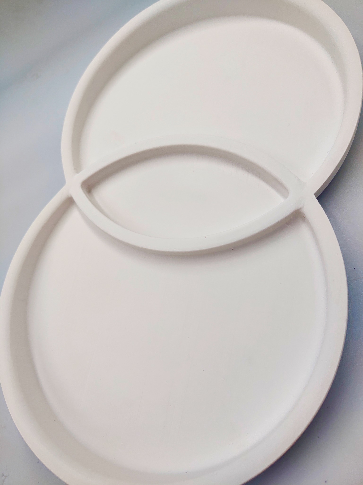 Białe, okrągłe formy z tworzywa sztucznego ułożone jedna na drugiej, z elementem łączącym w kształcie elipsy. Gładka powierzchnia, delikatne światło, minimalistyczny design.