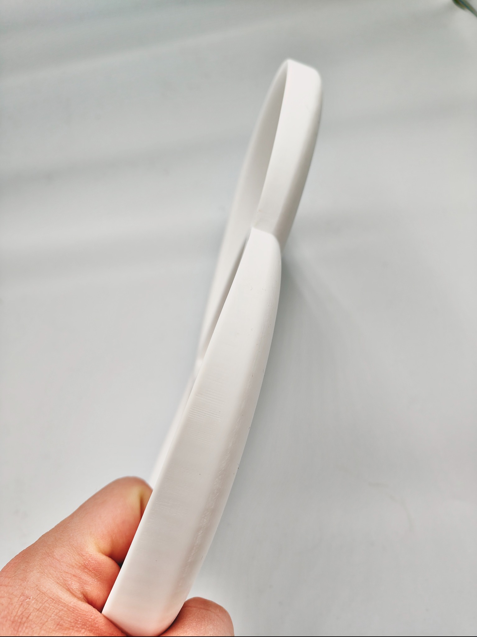 Biały, wydrukowany element w kształcie ósemki, trzymany w dłoni na tle jasnej powierzchni. Widoczne delikatne warstwy druku.