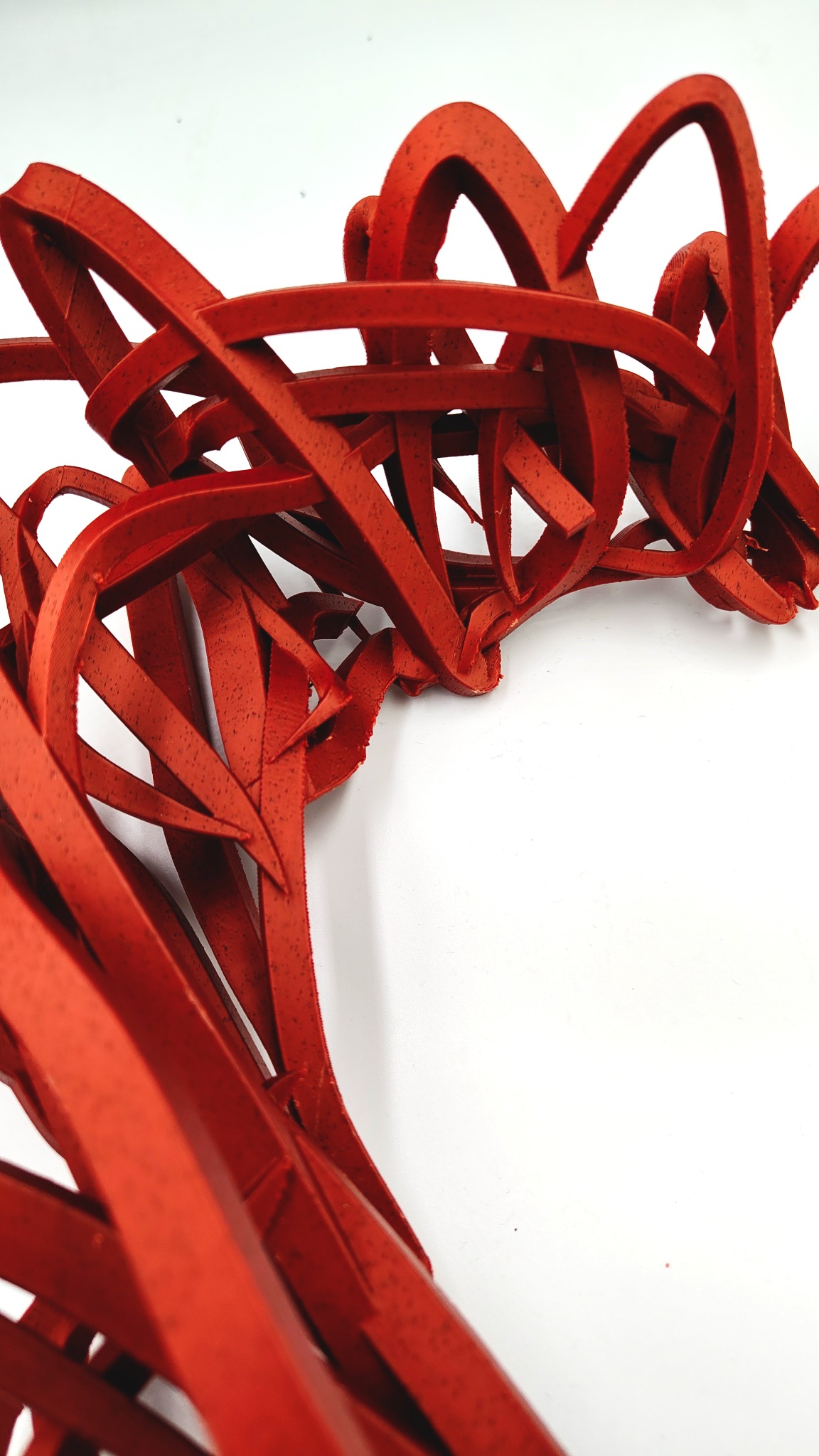 Czerwony, abstrakcyjny model z tworzywa sztucznego na białym tle. Widoczne warstwy i tekstura materiału. Nietypowy kształt, przypominający splątane wstążki.