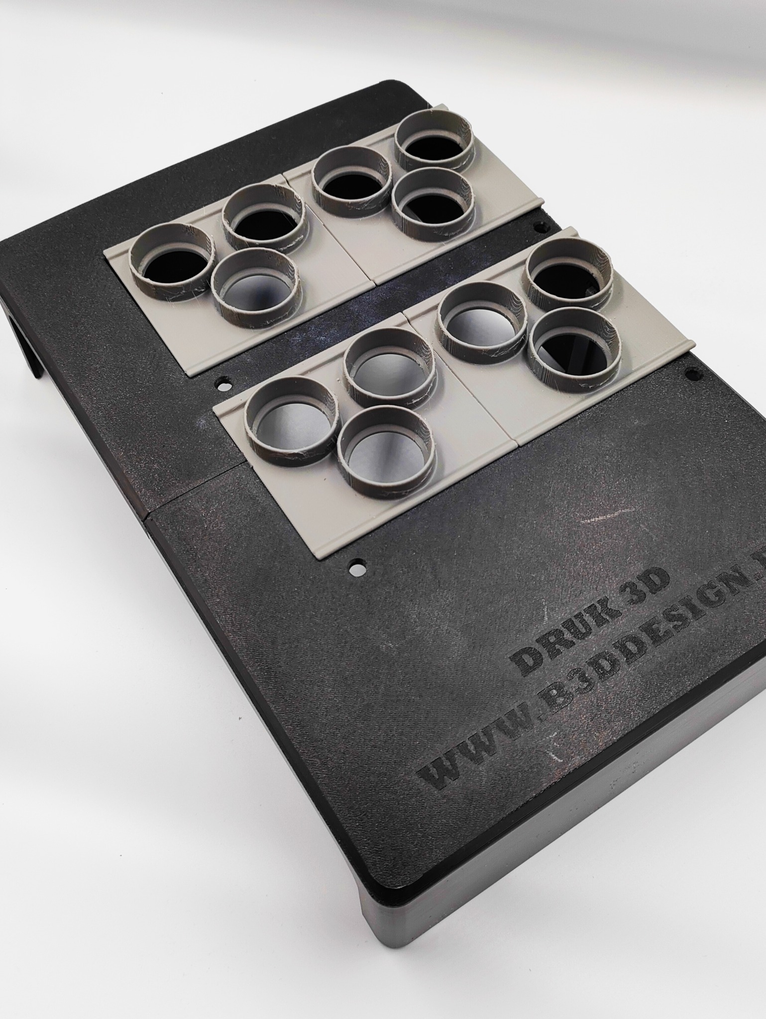 Druk 3D: Szare, okrągłe elementy na czarnej platformie z napisem 'DRUK 3D' i adresem strony. Widok z góry, oświetlenie studyjne.