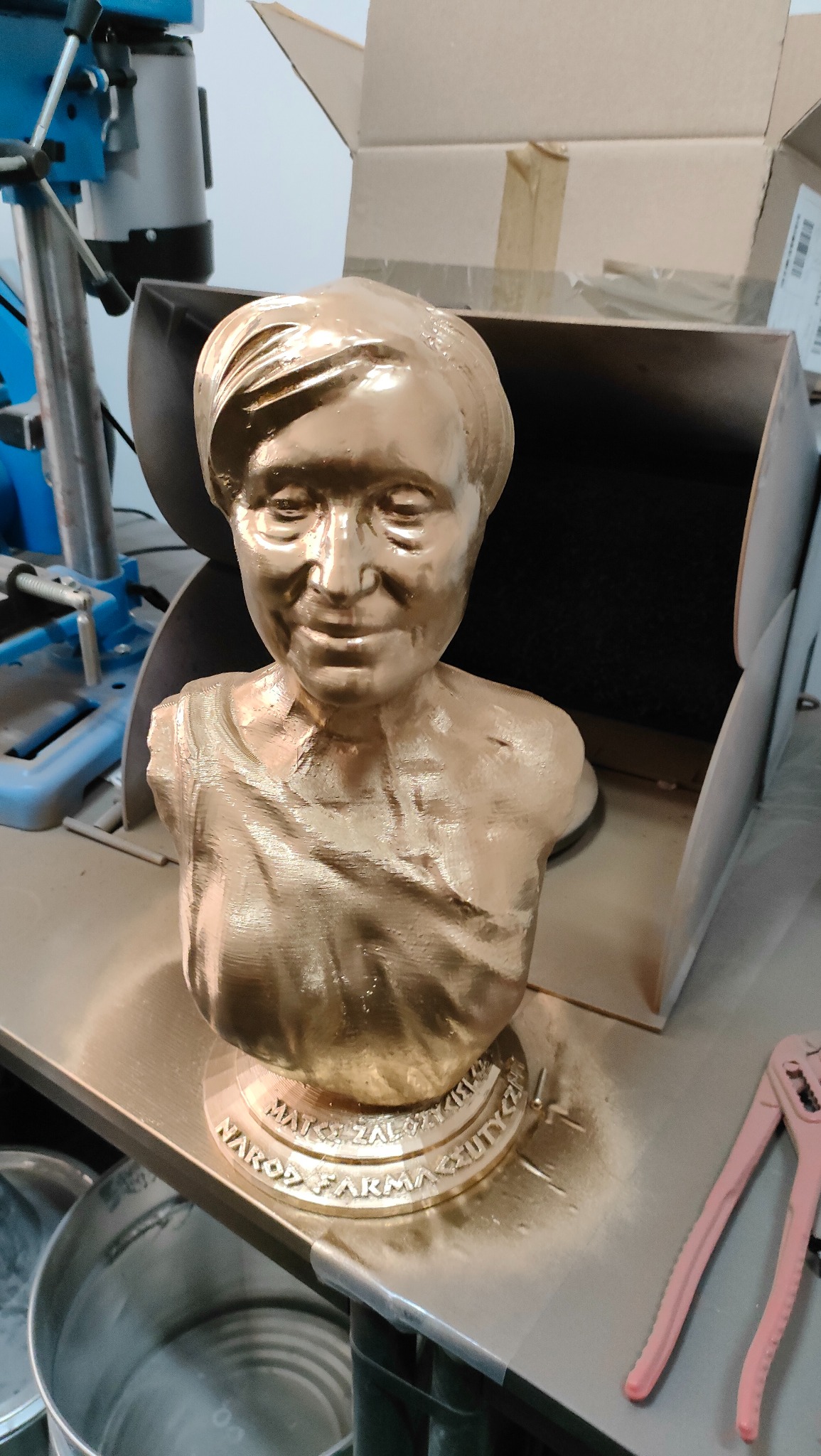 Złoty, pomalowany biust osoby, wydrukowany w 3D, z inskrypcją na podstawie. W tle widoczny warsztat z narzędziami, co sugeruje proces produkcyjny.