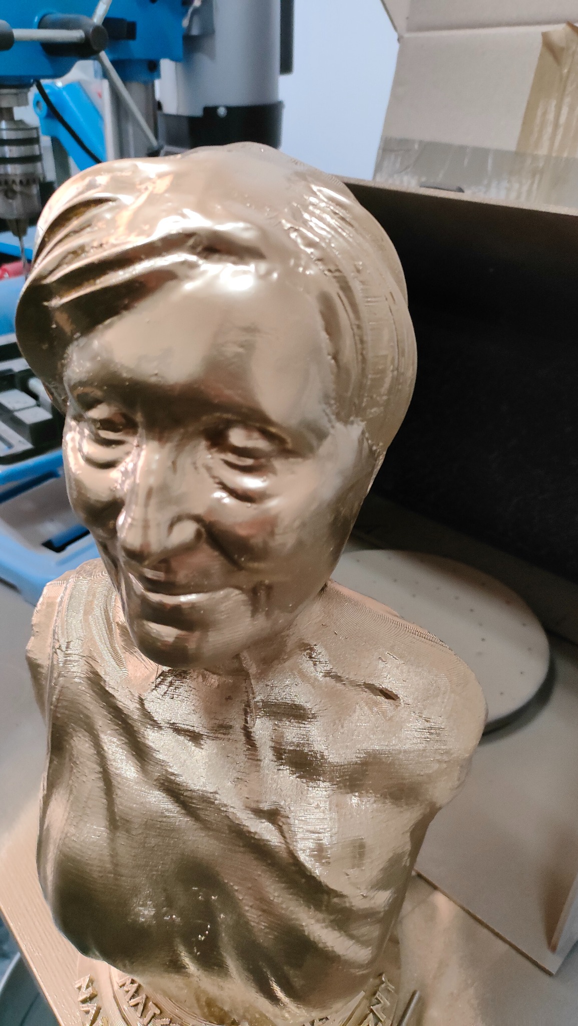 Złoty, pomalowany popiersie kobiety, wydrukowane w technologii FDM. Widoczne warstwy druku. W tle maszyna i karton.