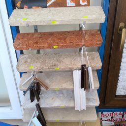 KRZYSZTOF MAZUR PROFIT PRZEDSIĘBIORSTWO BUDOWLANO-HANDLOWO-USŁUGOWE - Ekspozycja próbek okiennych i roletowych: blaty imitujące kamień, paleta kolorów drewna i wzorniki rolet w różnych odcieniach, prezentowane na metalowym regale.