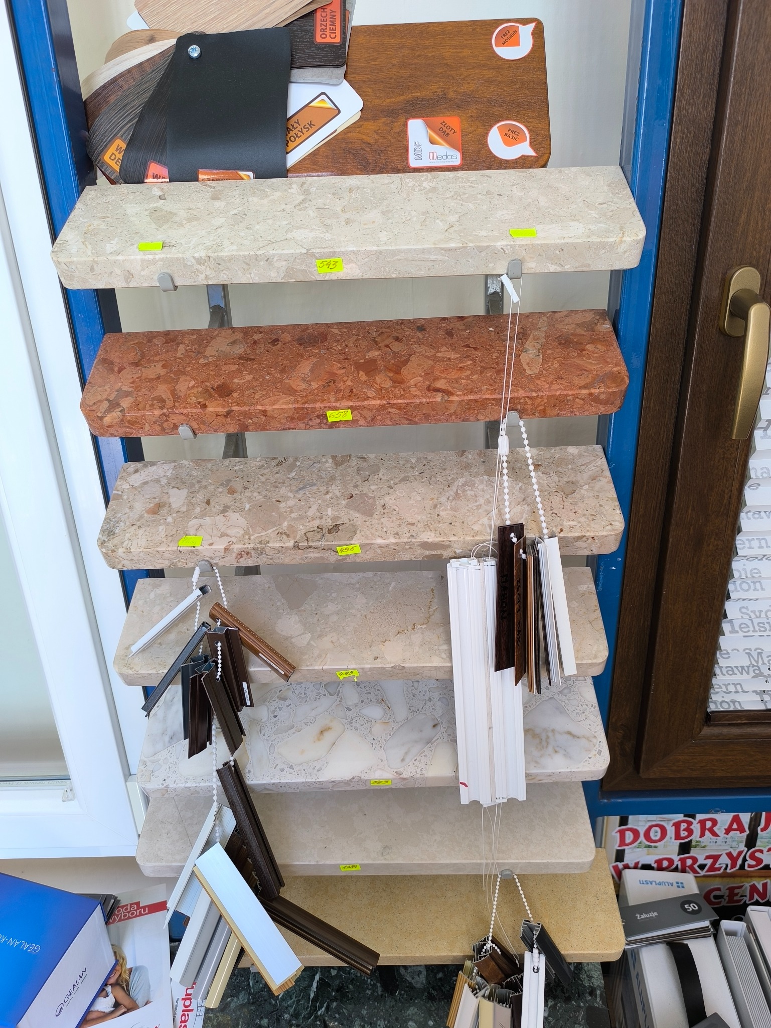 Ekspozycja próbek rolet wewnętrznych i okien PCV na półkach w salonie, prezentacja różnych kolorów i faktur materiałów wykończeniowych.