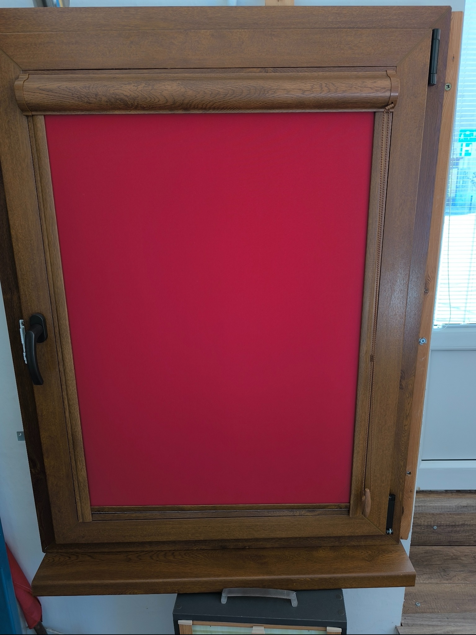 Drewniane okno z intensywnie czerwoną roletą zwiniętą u góry, widoczne detale mechanizmu łańcuszkowego i faktura drewna. Stonowana kolorystyka tła.