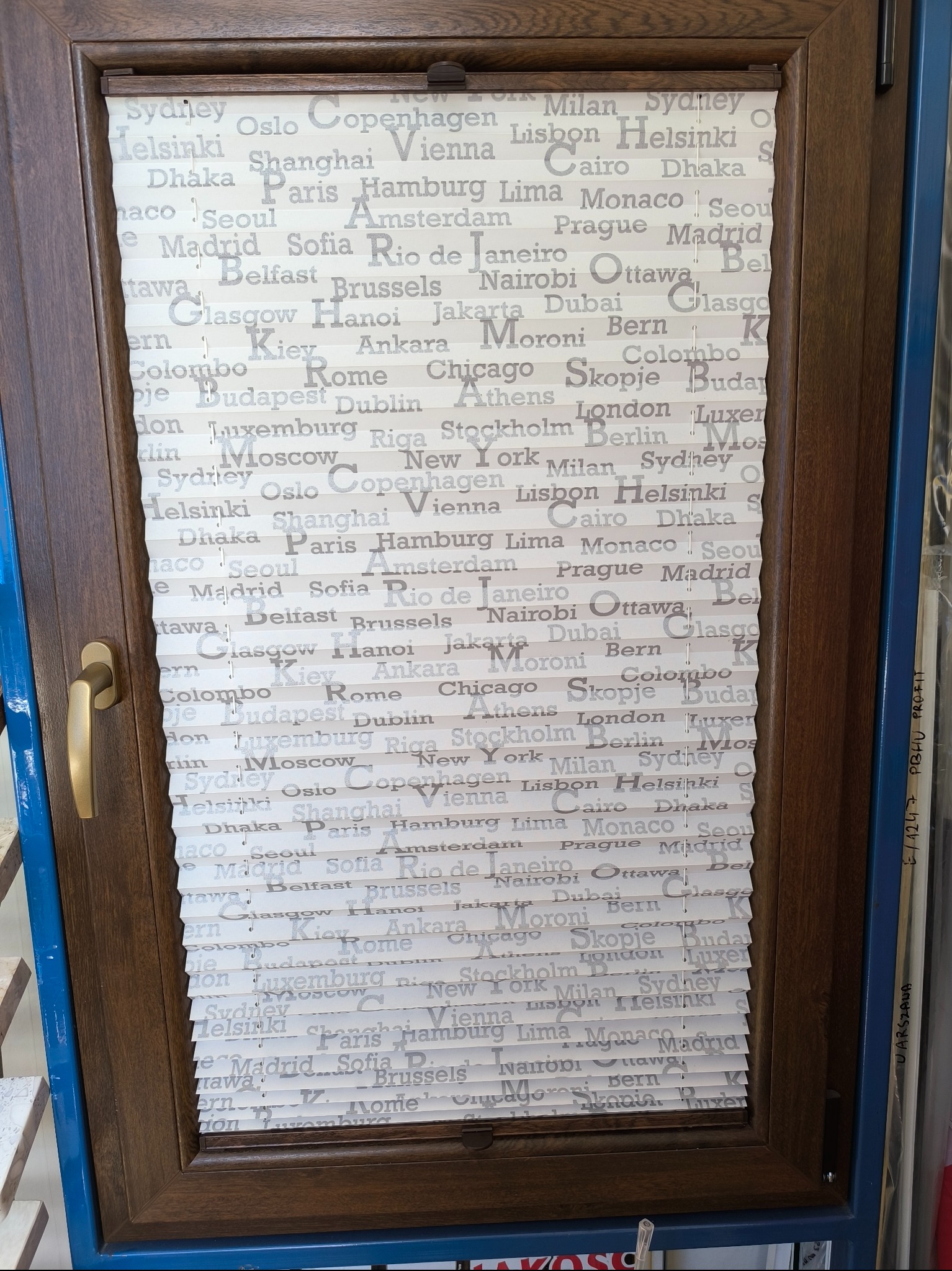 Brązowa rama okienna z plisowaną roletą wewnętrzną z motywem miast świata w odcieniach szarości. Roleta częściowo zasłonięta.