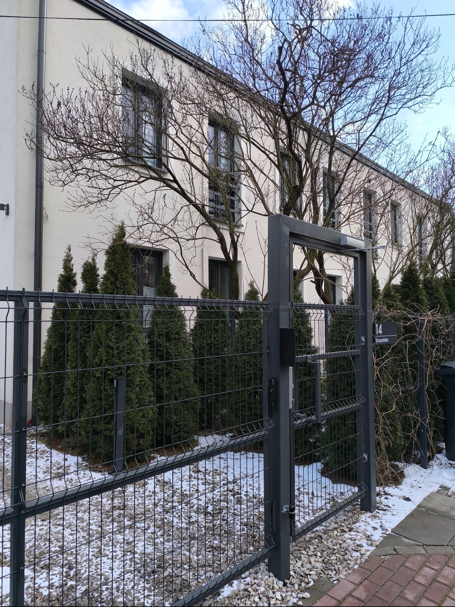 Nowoczesna, metalowa brama posesji z automatyką, widoczna z bliska na tle budynku z oknami i zielenią. Zima, lekko ośnieżone podłoże.