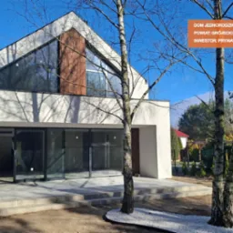 Nowoczesny dom jednorodzinny z białymi ścianami i drewnianymi elementami elewacji, widoczne duże przesuwne okna tarasowe i fragment zagospodarowanego terenu z drzewami.