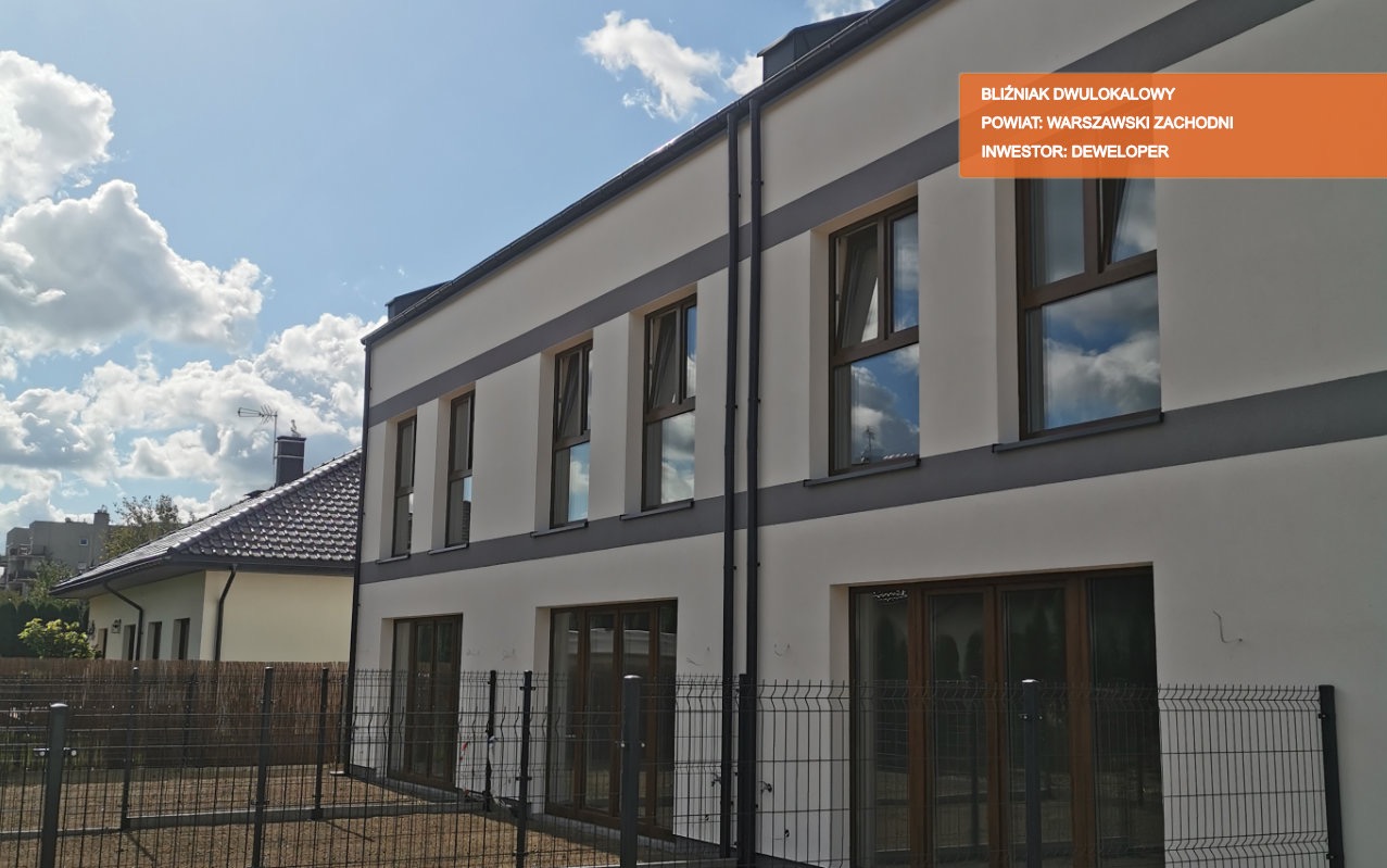 Dwukondygnacyjny budynek bliźniaczy z białymi ścianami, szarymi akcentami i brązowymi oknami, widoczny z zewnątrz za czarnym ogrodzeniem z siatki, sąsiedni dom z brązowym dachem w tle.