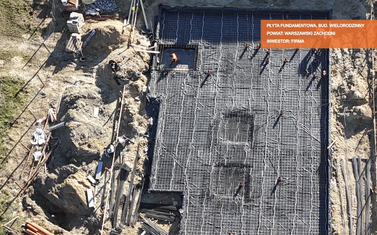 Z góry widoczna płyta fundamentowa zbrojona siatką stalową, przygotowana do wylania betonu na budowie wielorodzinnej w powiecie warszawskim zachodnim; pracownik w pomarańczowej kamizelce w wykopie.