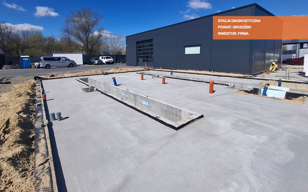 Wylany betonowy fundament pod budynek stacji diagnostycznej, widoczne pionowe rury instalacyjne i izolacja przeciwwilgociowa, w tle budynek z bramą segmentową i zaparkowane samochody.