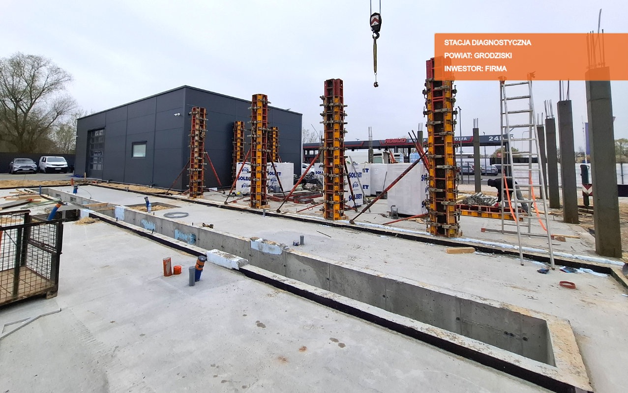 Budowa stacji diagnostycznej: widok na fundamenty i szalunki kolumn, z widocznym budynkiem stacji w tle oraz dźwigiem w górnej części kadru.