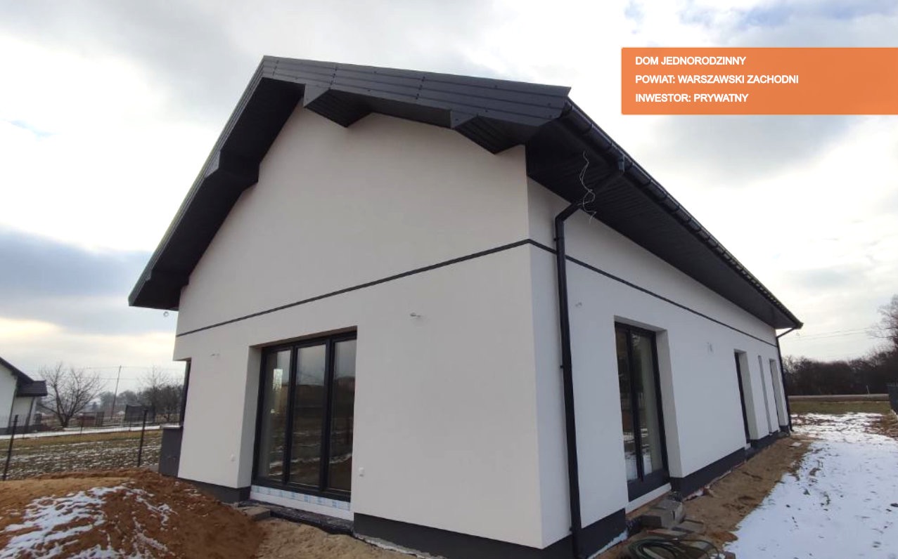 Narożne ujęcie z zewnątrz, prezentujące nowoczesny dom jednorodzinny z białymi ścianami, czarnym dachem i ramami okiennymi, z widocznymi śladami śniegu na ziemi.