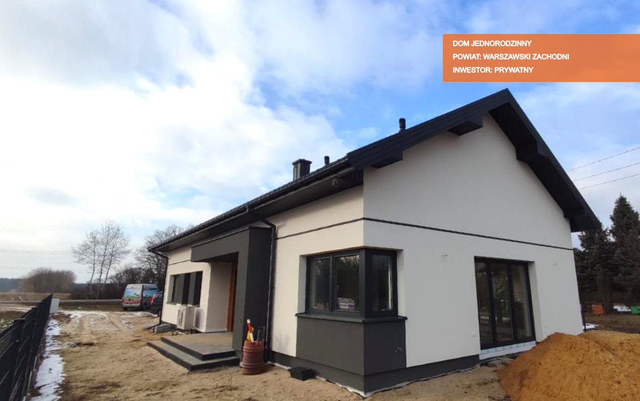 Nowoczesny dom jednorodzinny z białą elewacją i antracytowymi detalami, widok z zewnątrz na etapie wykończenia terenu, z widoczną kupką piasku i częściowo zasypanym fundamentem.