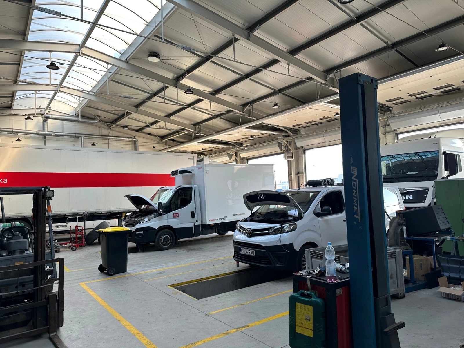 Wnętrze warsztatu samochodowego z dwoma dostawczymi pojazdami z otwartymi maskami nad kanałami diagnostycznymi, w tle zaparkowane ciężarówki, widoczny podnośnik kolumnowy, wózek narzędziowy i inne...