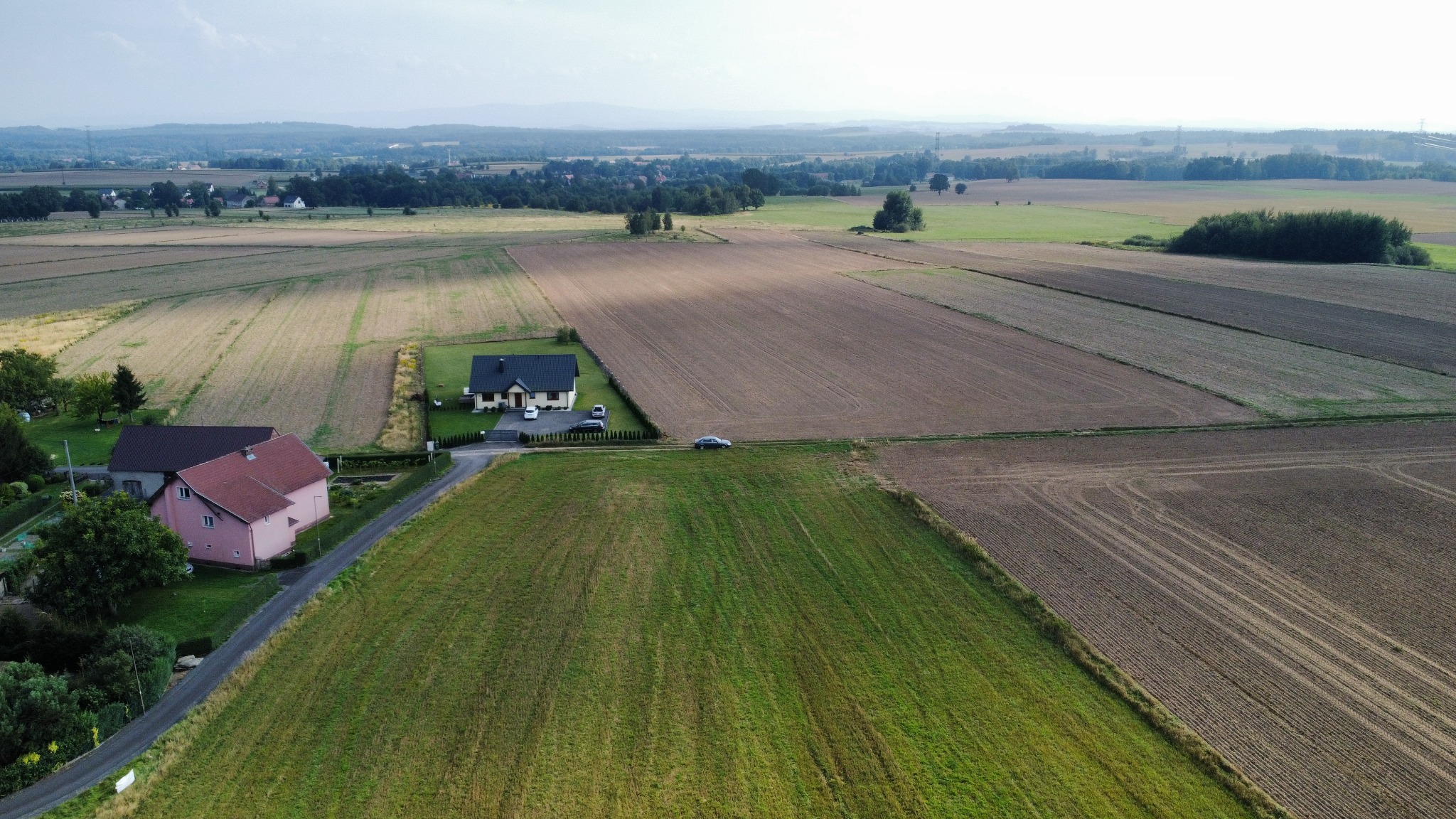 Panoramiczny widok z lotu ptaka na krajobraz rolniczy z domami, polami uprawnymi w różnych stadiach wzrostu, samochodem na drodze i odległymi wzgórzami na horyzoncie.