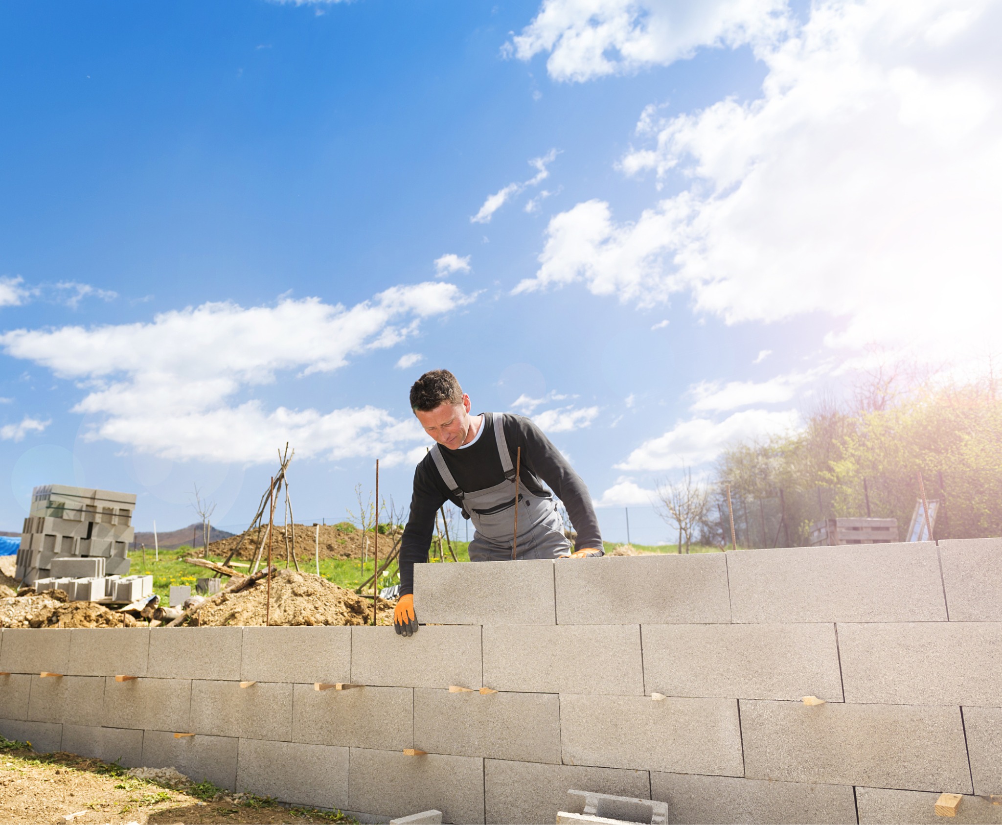 Murarz w szarym kombinezonie i pomarańczowych rękawicach układa ścianę z szarych bloczków betonowych na tle błękitnego nieba i zieleni.