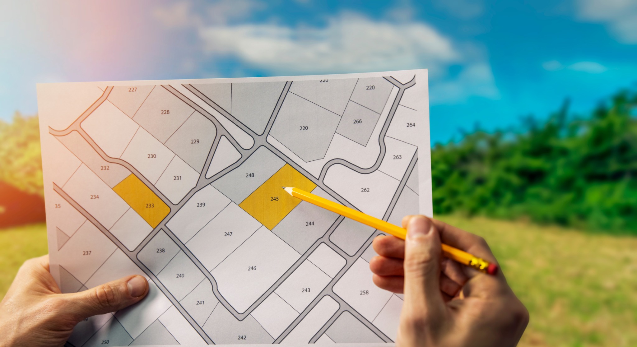 Osoba trzyma plan działek z zaznaczonymi numerami 233 i 245, wskazując ołówkiem na działkę 245, na tle błękitnego nieba i zieleni.