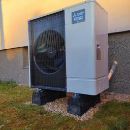 KPM Patrycja Centner - Zewnętrzna jednostka pompy ciepła Mitsubishi Zubadan R32 zamontowana na gumowych podstawach na trawniku przy elewacji budynku.