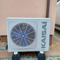 KPM Patrycja Centner - Zewnętrzna jednostka klimatyzacji Kaisai, zamontowana na czarnych gumowych podstawach, ustawiona na betonowej kostce brukowej przed beżową ścianą budynku z rynną spustową.