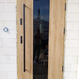 KPM Patrycja Centner - Montaż nowoczesnych drzwi wejściowych z okładziną drewnopodobną i czarnym szkłem, widoczna ościeżnica uszczelniona pianką montażową, ściana z białych bloczków.