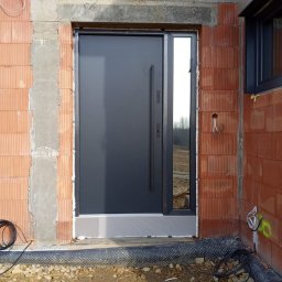 KPM Patrycja Centner - Nowoczesne, antracytowe drzwi wejściowe z bocznym przeszkleniem zamontowane w surowym murze z czerwonej cegły, widoczne przewody elektryczne i fragment okna po prawej stronie.