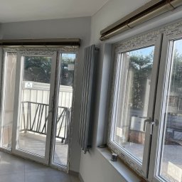 KPM Patrycja Centner - Szary grzejnik panelowy zamontowany na ścianie obok białych okien PCV z widokiem na zewnątrz, z balkonem i zielenią, w jasnym pomieszczeniu z szarą podłogą.