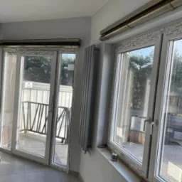 Szary grzejnik panelowy zamontowany na ścianie obok białych okien PCV z widokiem na zewnątrz, z balkonem i zielenią, w jasnym pomieszczeniu z szarą podłogą.