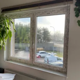 Nowe okno PCV z roletami wewnętrznymi, widok na podjazd z samochodami i drzewa w tle.