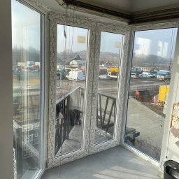 KPM Patrycja Centner - Zabudowany balkon z białymi oknami PCV marki Schuco, widok na plac budowy z kontenerami i maszynami, szare płytki na podłodze, kamera monitoringu na suficie.