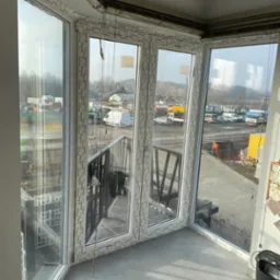 Zabudowany balkon z białymi oknami PCV marki Schuco, widok na plac budowy z kontenerami i maszynami, szare płytki na podłodze, kamera monitoringu na suficie.