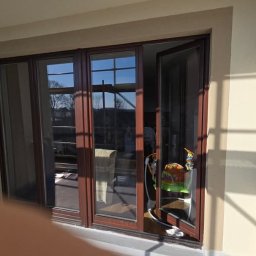 KPM Patrycja Centner - Nowe, brązowe okno balkonowe trzyskrzydłowe, częściowo otwarte, z widokiem na zewnątrz, z jasnymi refleksami słonecznymi na ścianie i podłodze.