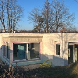 KPM Patrycja Centner - Surowy stan domu z bloczków, widoczne okna z ociepleniem, żółty wąż leży na ziemi, w tle zabudowa miejska z wieżą kościelną.