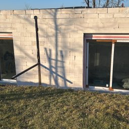 KPM Patrycja Centner - Ściana fundamentowa z bloczków betonu komórkowego, z dwoma oknami z brązowymi ramami, widoczne ocieplenie pionowe po bokach okien, cień drabiny na ścianie, trawa na pierwszym planie.