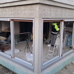 KPM Patrycja Centner - Montaż narożnego okna panoramicznego w surowym stanie, z widocznym pracownikiem na drabinie, laserem na statywie i materiałami budowlanymi wewnątrz.