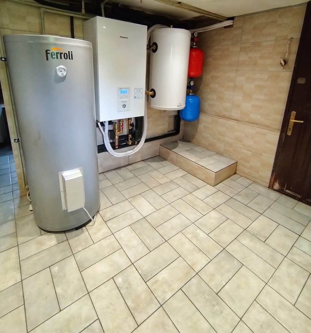 Kotłownia z zainstalowanym piecem gazowym Panasonic, zasobnikiem Ferroli, zbiornikami wyrównawczymi w kolorach czerwonym i niebieskim, wykończona kafelkami w odcieniach beżu.