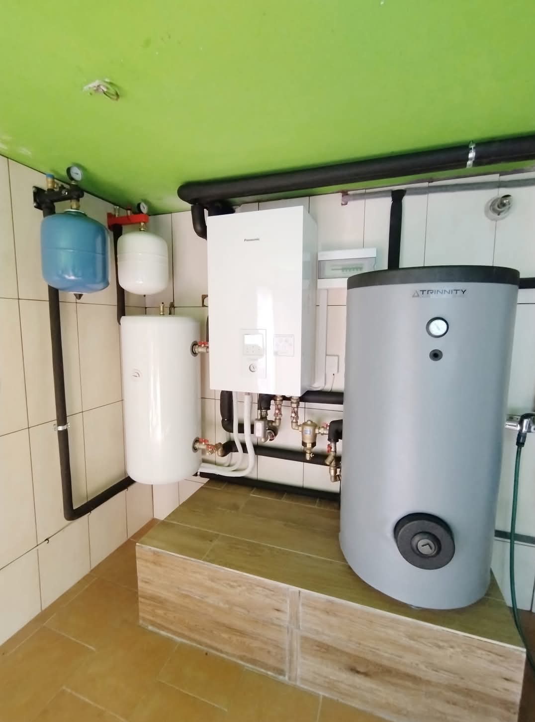 Zestaw urządzeń grzewczych: wiszący piec gazowy Panasonic, zbiornik buforowy Trinity, naczynia przeponowe w instalacji centralnego ogrzewania, rury instalacyjne na tle białych płytek i zielonego...