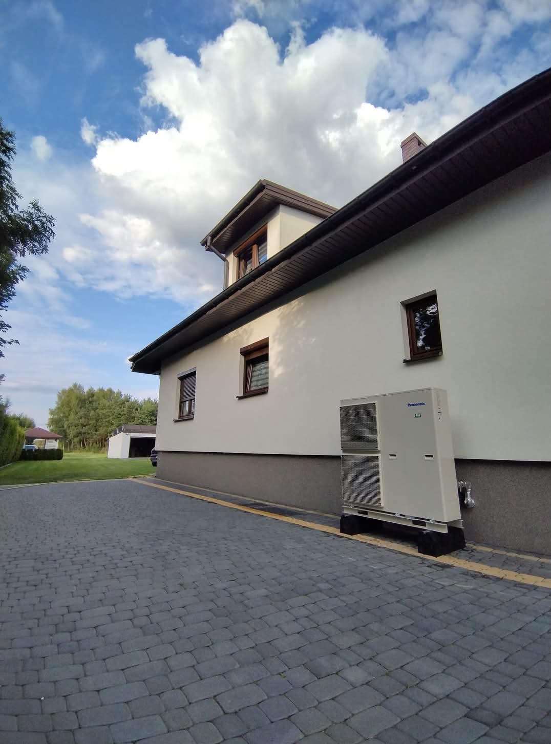 Biała pompa ciepła Panasonic zamontowana na zewnątrz domu jednorodzinnego z szarą elewacją i brukowanym podjazdem w słoneczny dzień.