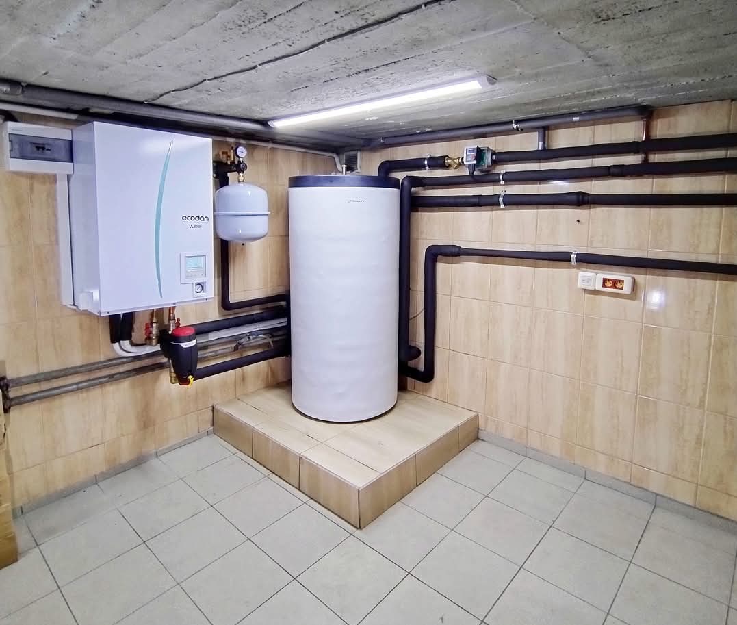 Biały zbiornik buforowy i jednostka grzewcza Ecodan w kotłowni z wykafelkowanymi ścianami i podłogą, orurowanie w izolacji termicznej.