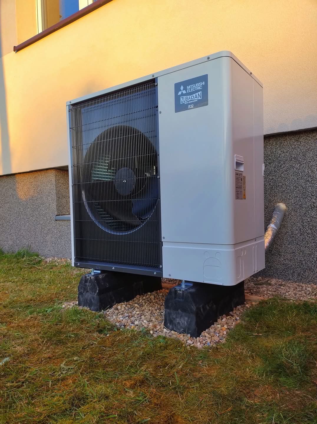 Zewnętrzna jednostka pompy ciepła Mitsubishi Zubadan R32 zamontowana na gumowych podstawach na trawniku przy elewacji budynku.